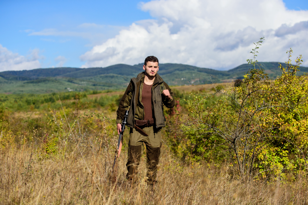 Man walking in long grass wearing camouflage suit with a rifle by his side.